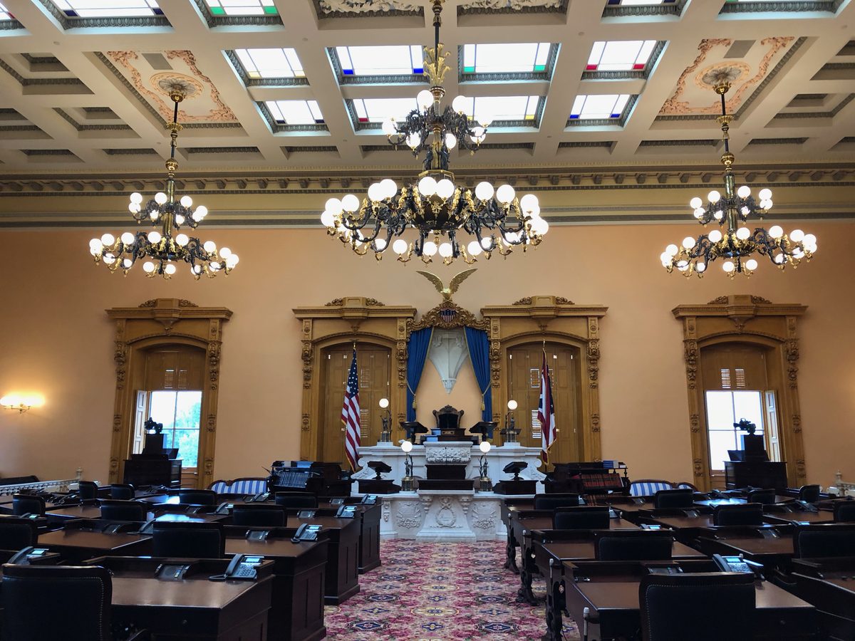 Empty Ohio Senate chamber with dark wood desks, blue drapery, flags, and chandeliers.